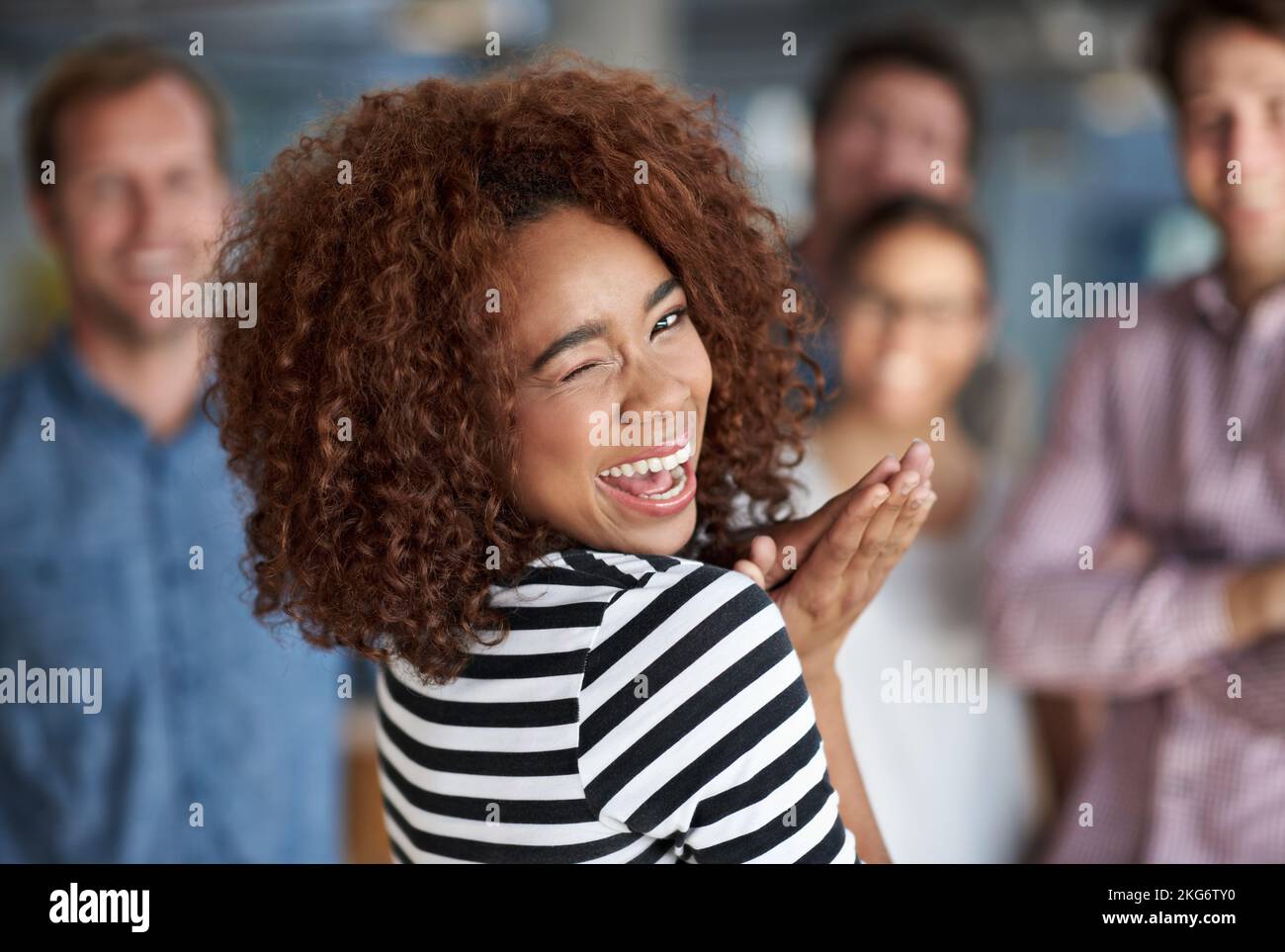 Shes the life of the office. Young woman smiling cheekily at the camera with colleagues in the background. Stock Photo