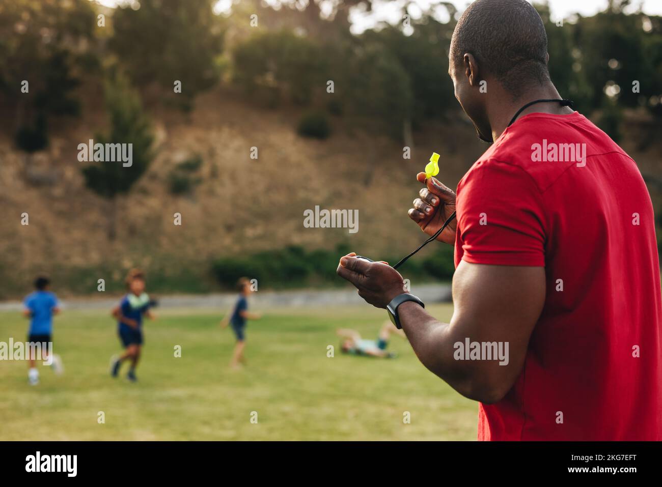 Coach holding a whistle during a PE session with school kids. Man training a group of elementary school children in a school ground. Sports and recrea Stock Photo