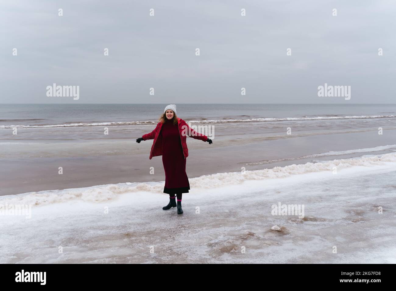 A joyful woman in a red jacket and a dress by the sea in winter. Stock Photo