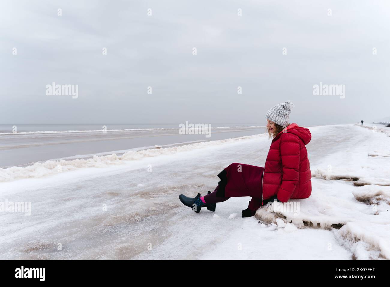 A cheerful woman in a red jacket and a warm dress sits on the shore of the winter sea. Stock Photo