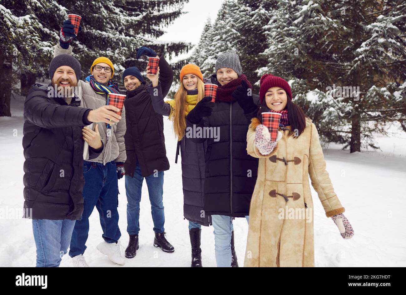 Cheerful friends during winter walk raise up paper cups with hot drink cheering and greeting you. Stock Photo