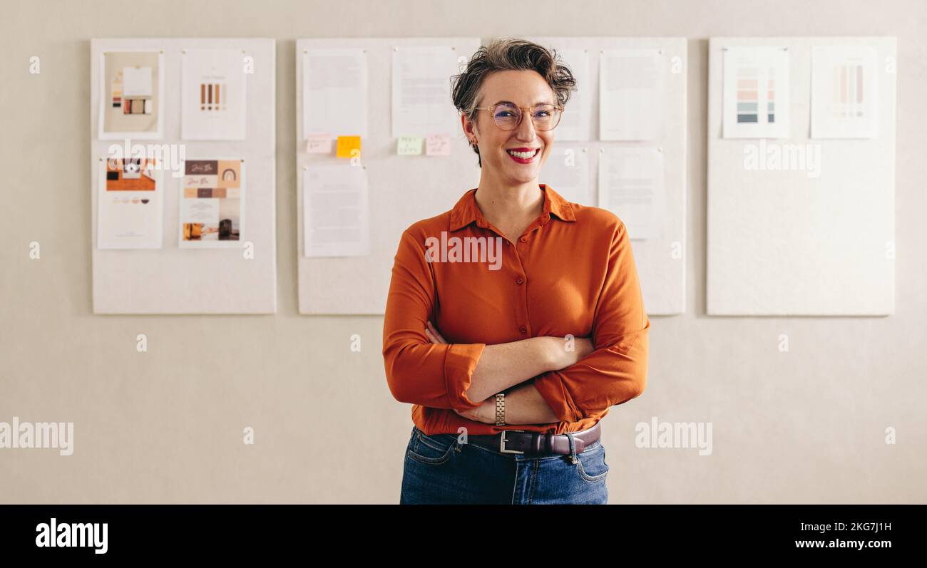 Portrait of a cheerful female designer smiling at the camera while standing with her arms crossed. Mature businesswoman with eyeglasses working in a c Stock Photo