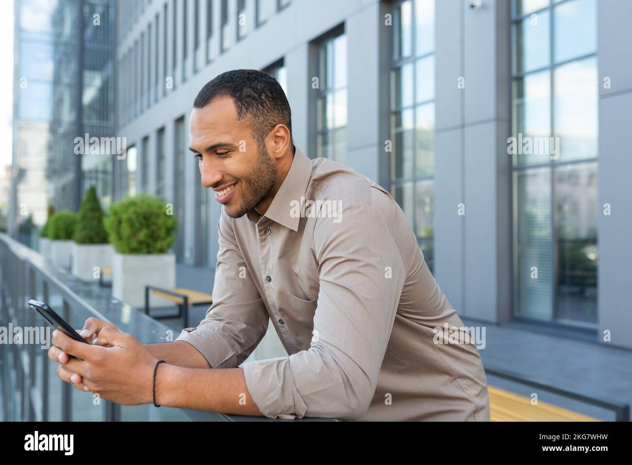 A young handsome Latin American man is standing on the veranda of the office center, holding a phone in his hands, typing a message, writing a text, using it. Stock Photo