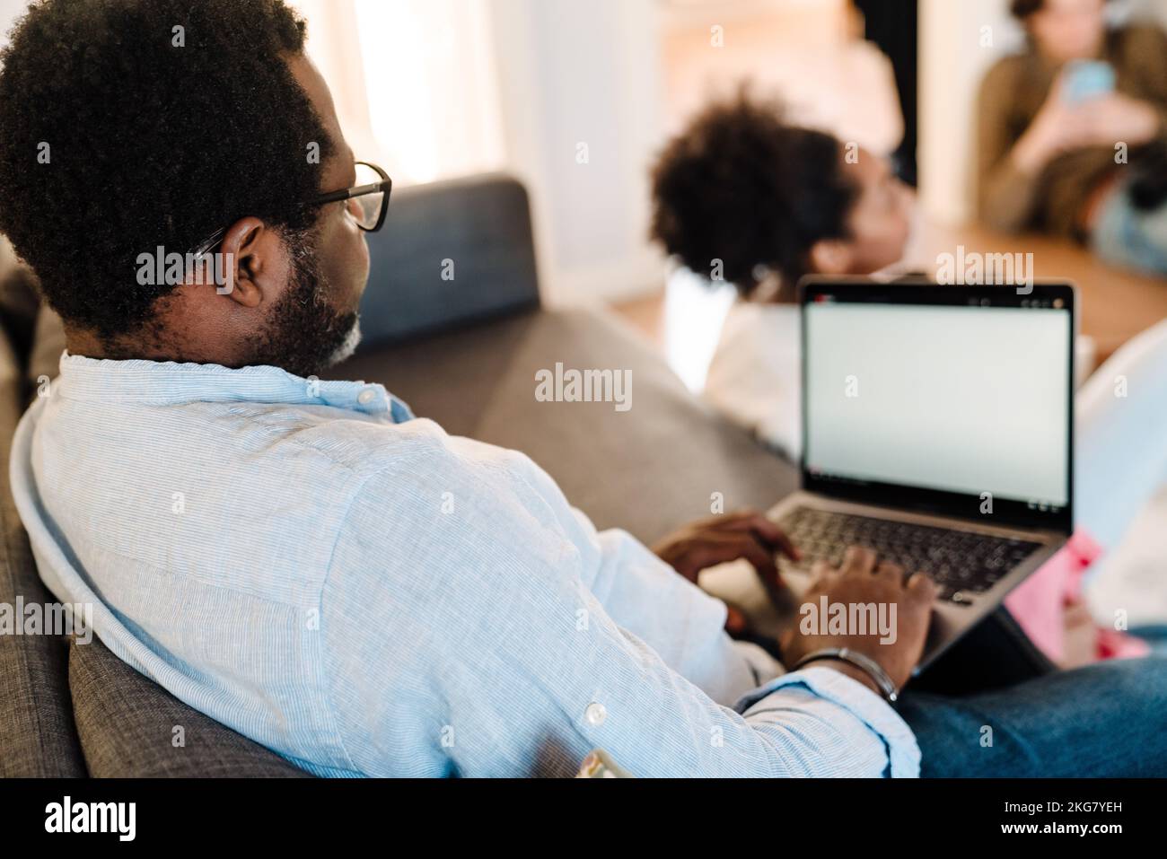 African american man using laptop while sitting on sofa with family at ...