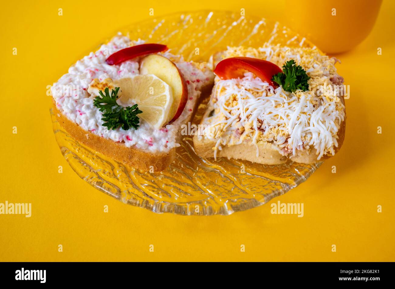 2 sandwiches on glass plate on yellow background. Right with spread with cheese, boiled egg, ham and apple, left with lobster spread, apple and lemon. Stock Photo