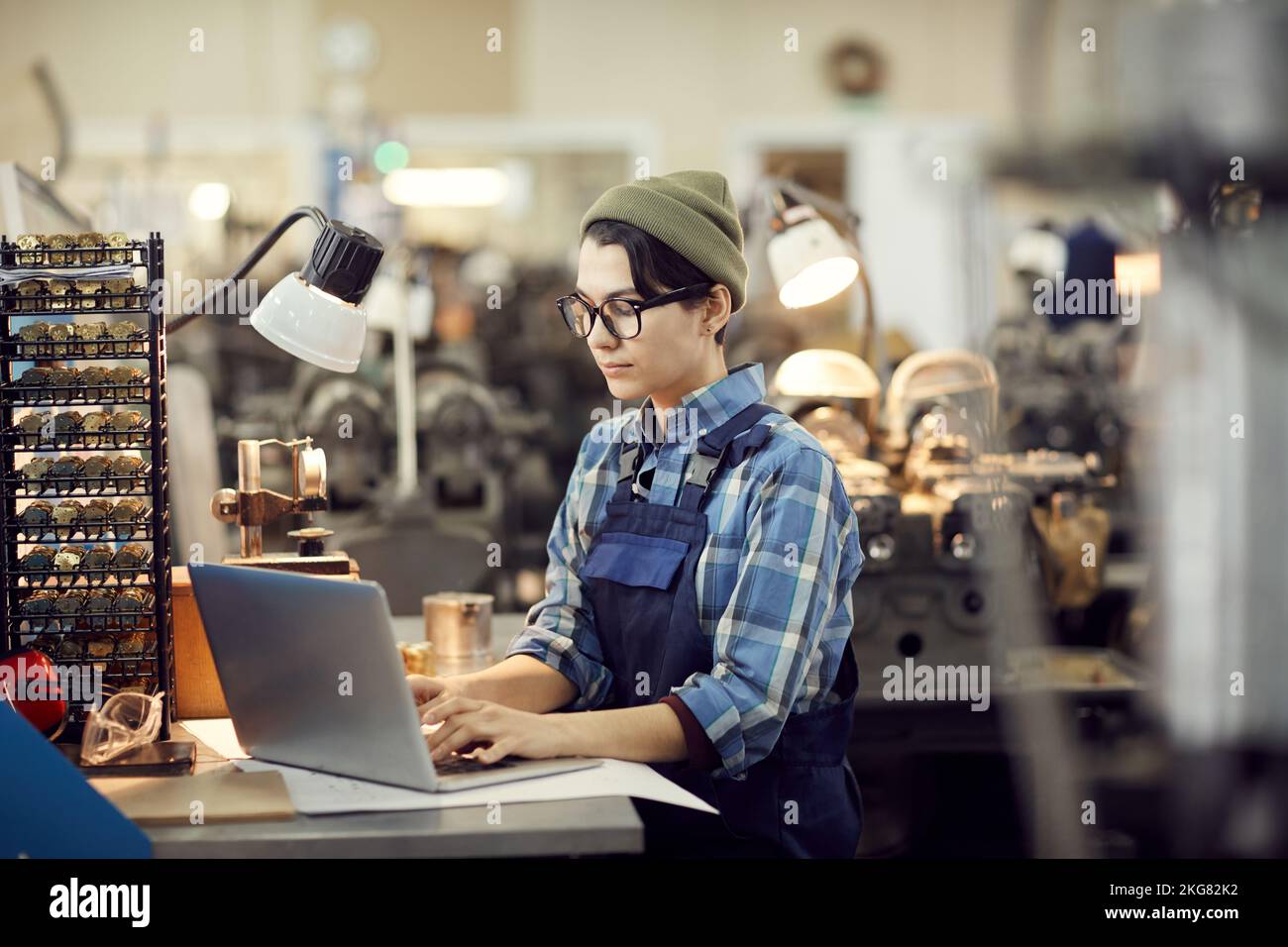Serious skilled factory girl in eyeglasses sitting at table and making records on laptop while working at watch plant Stock Photo