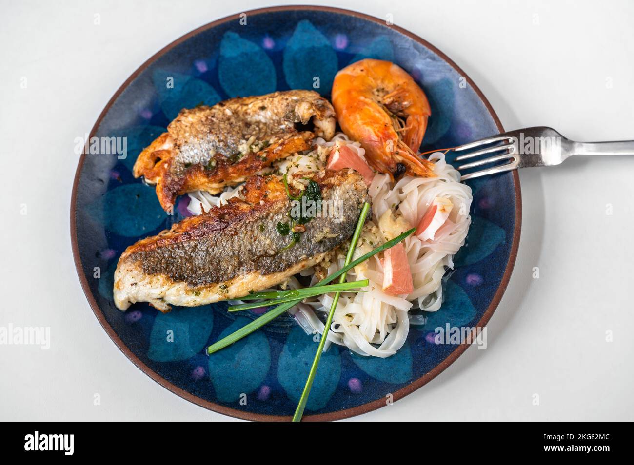 Fried sea bream fillet and shrimp with glass noodle on blue decorative plate with fork on white background. Stock Photo