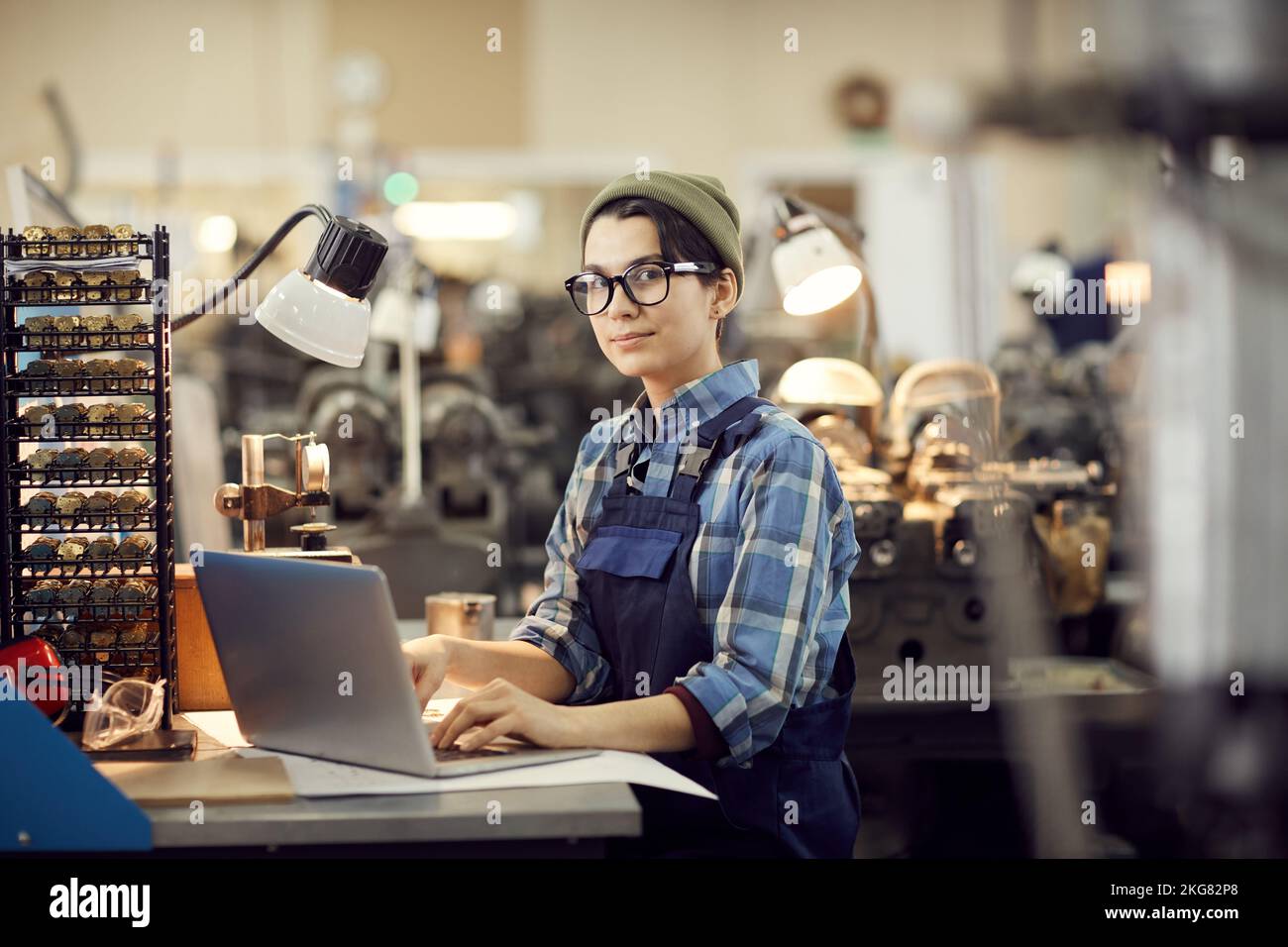 Portrait of content hipster female worker in eyeglasses sitting at desk and using laptop while composing manual for assembling wristwatch Stock Photo