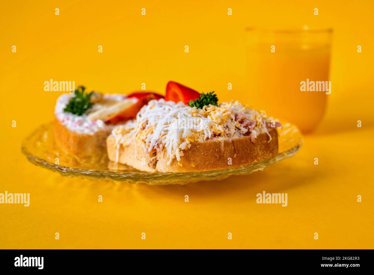 2 sandwiches on glass plate, glass of juice on yellow background. Right sandwich with spread with cheese, boiled egg, ham and apple, left with lobster Stock Photo