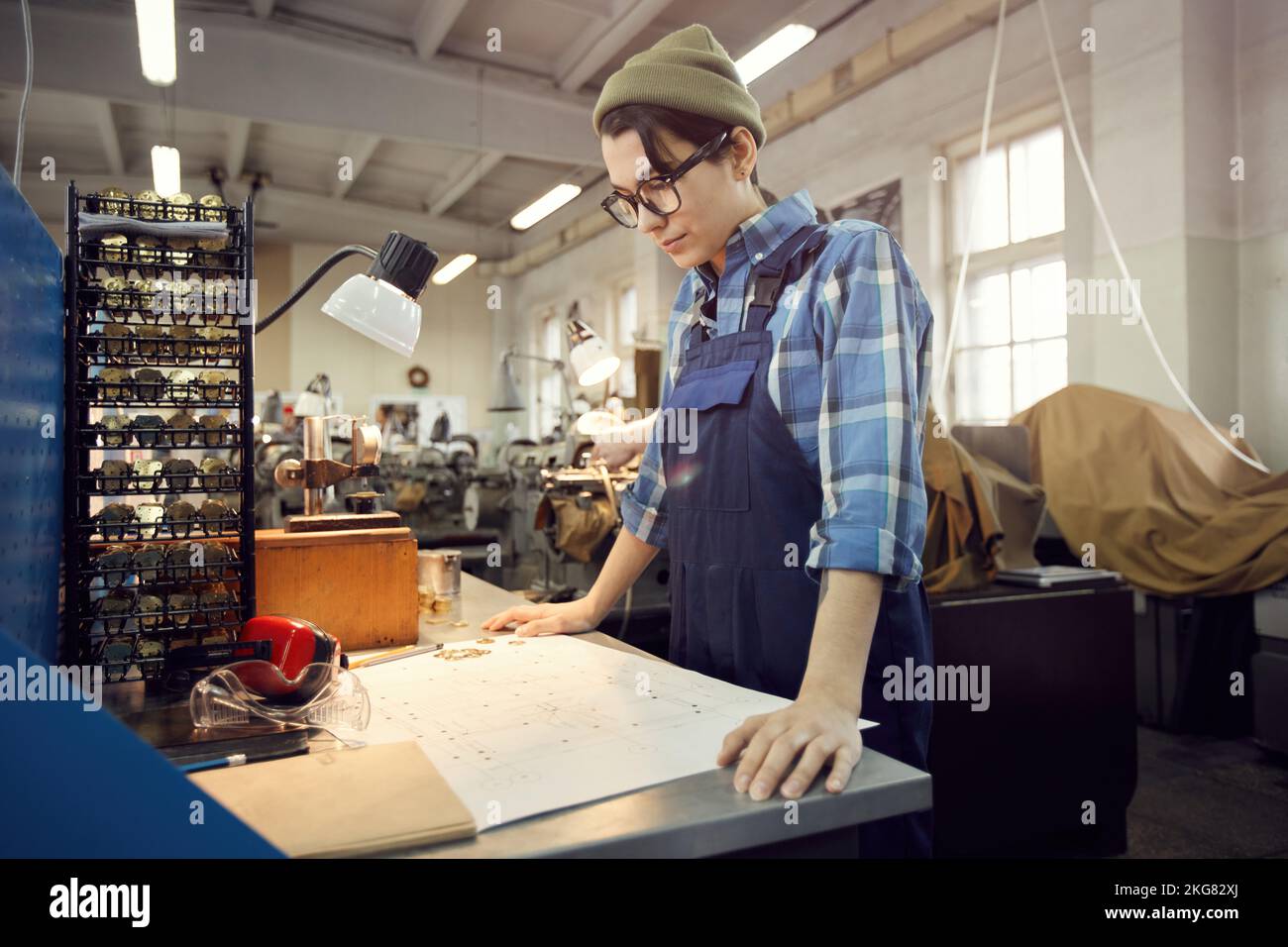 Serious concentrated young woman in workwear standing at table and watching assembly plan of watch movement Stock Photo