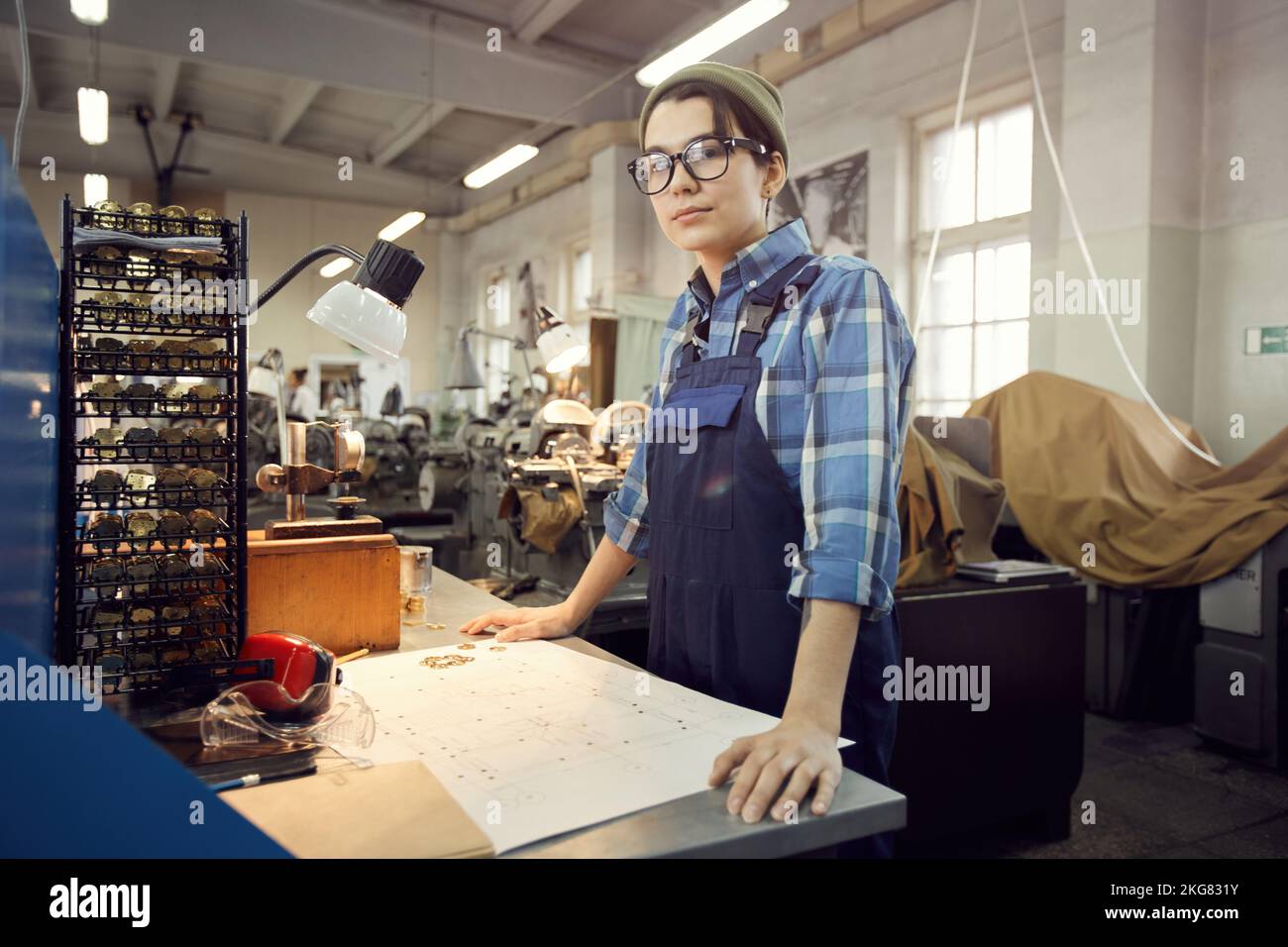 Portrait of serious female production worker in hipster hat standing at desk and assembling wristwatch movement according to scheme Stock Photo