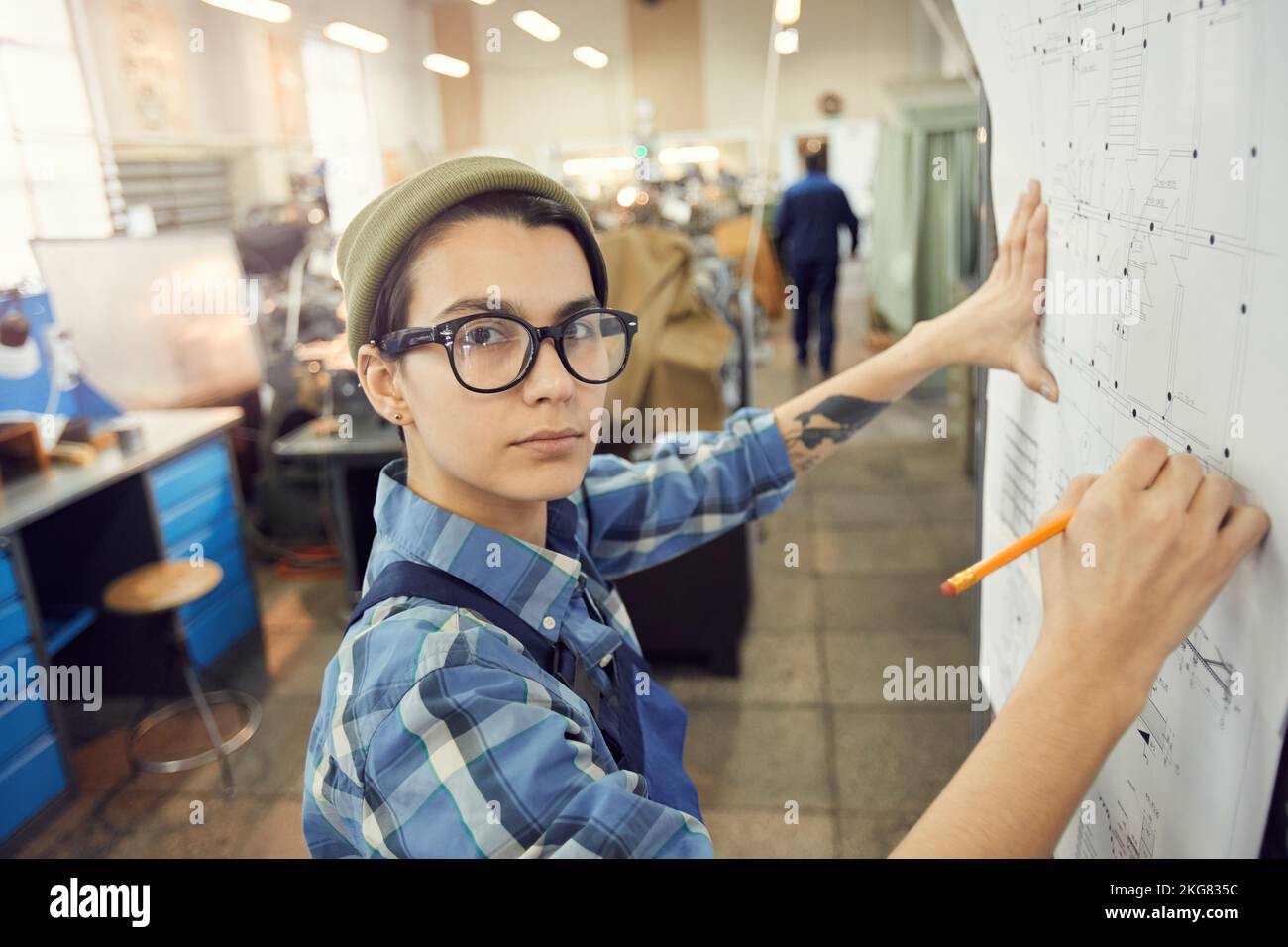 Portrait of serious skilled factory worker in eyeglasses standing at wall in factory shop and analyzing engineering blueprint Stock Photo
