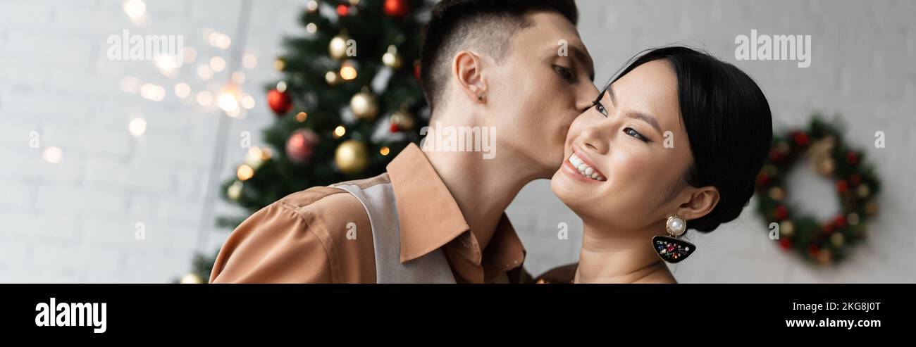 young man kissing cheerful asian wife while celebrating Christmas at home, banner Stock Photo