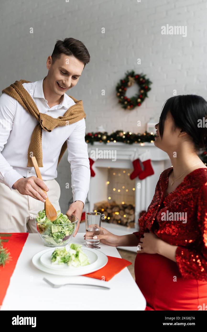 pregnant asian woman sitting with glass of water near smiling husband holding bowl with vegetable salad Stock Photo