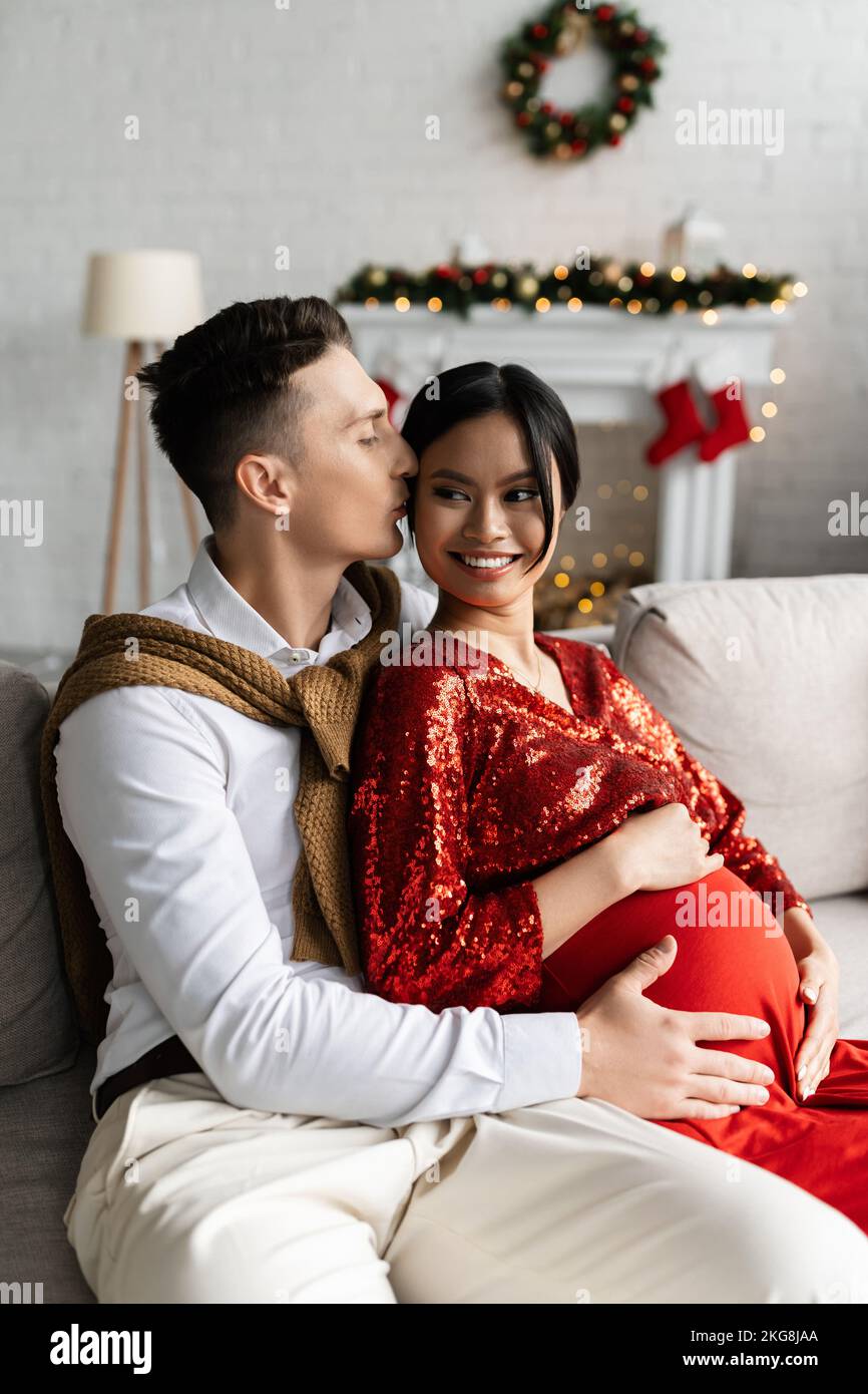 man kissing pregnant asian woman and hugging her belly during Christmas celebration Stock Photo