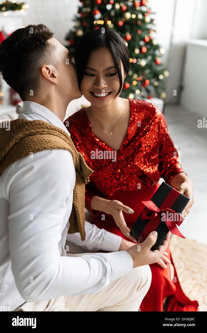 young man holding Christmas present and kissing pregnant asian woman in elegant clothes Stock Photo