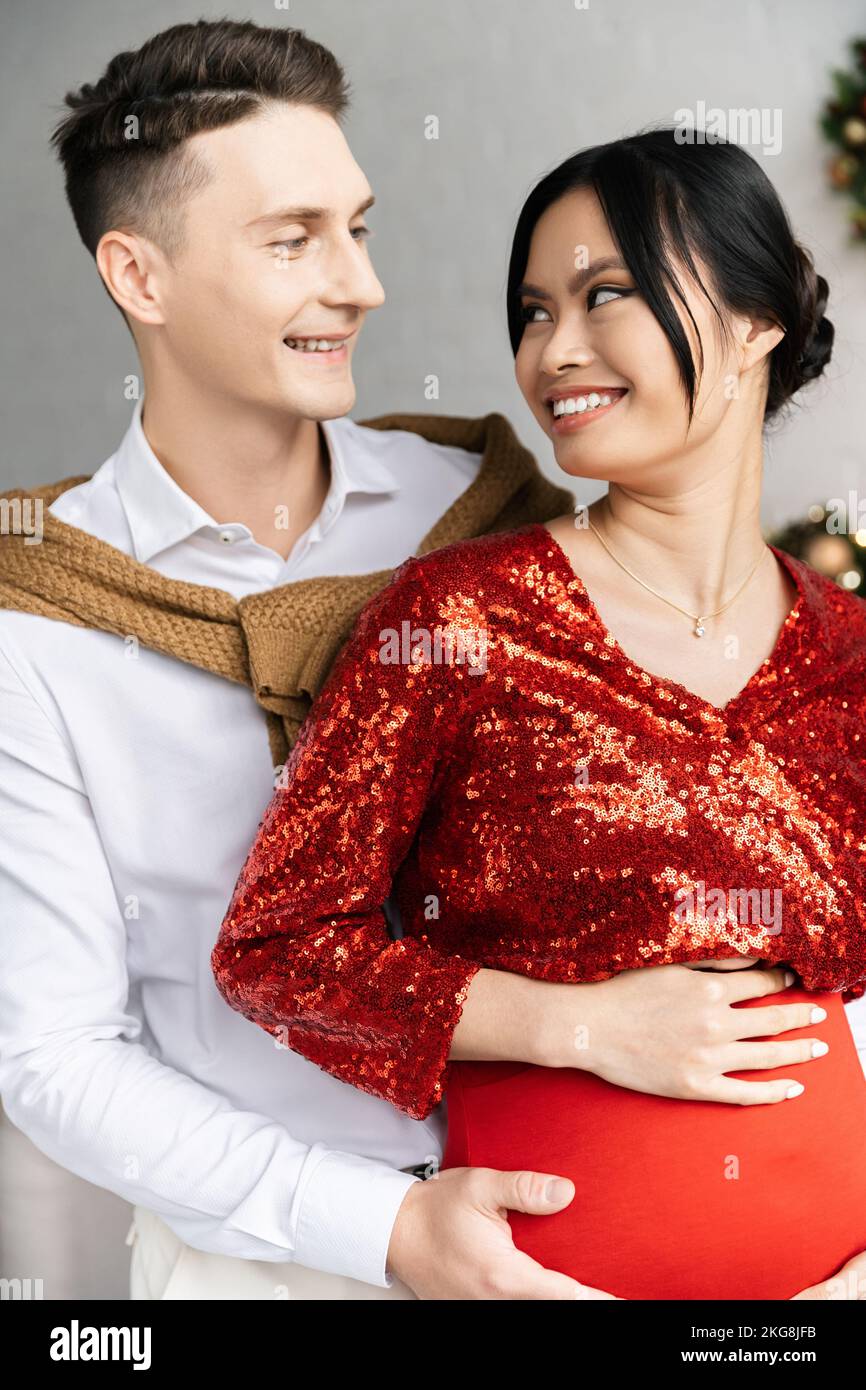 happy man and pregnant asian woman in festive clothes smiling at each other Stock Photo