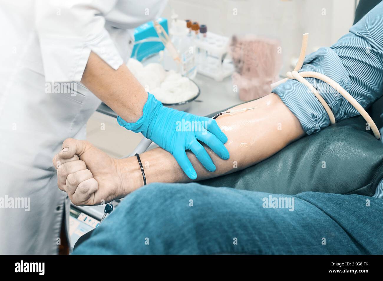 Medical worker in gloves and gown takes blood from man's vein. Donor sits in chair and donates blood. Background. Authentic scene from medical clinic. Topic of donation.. Stock Photo