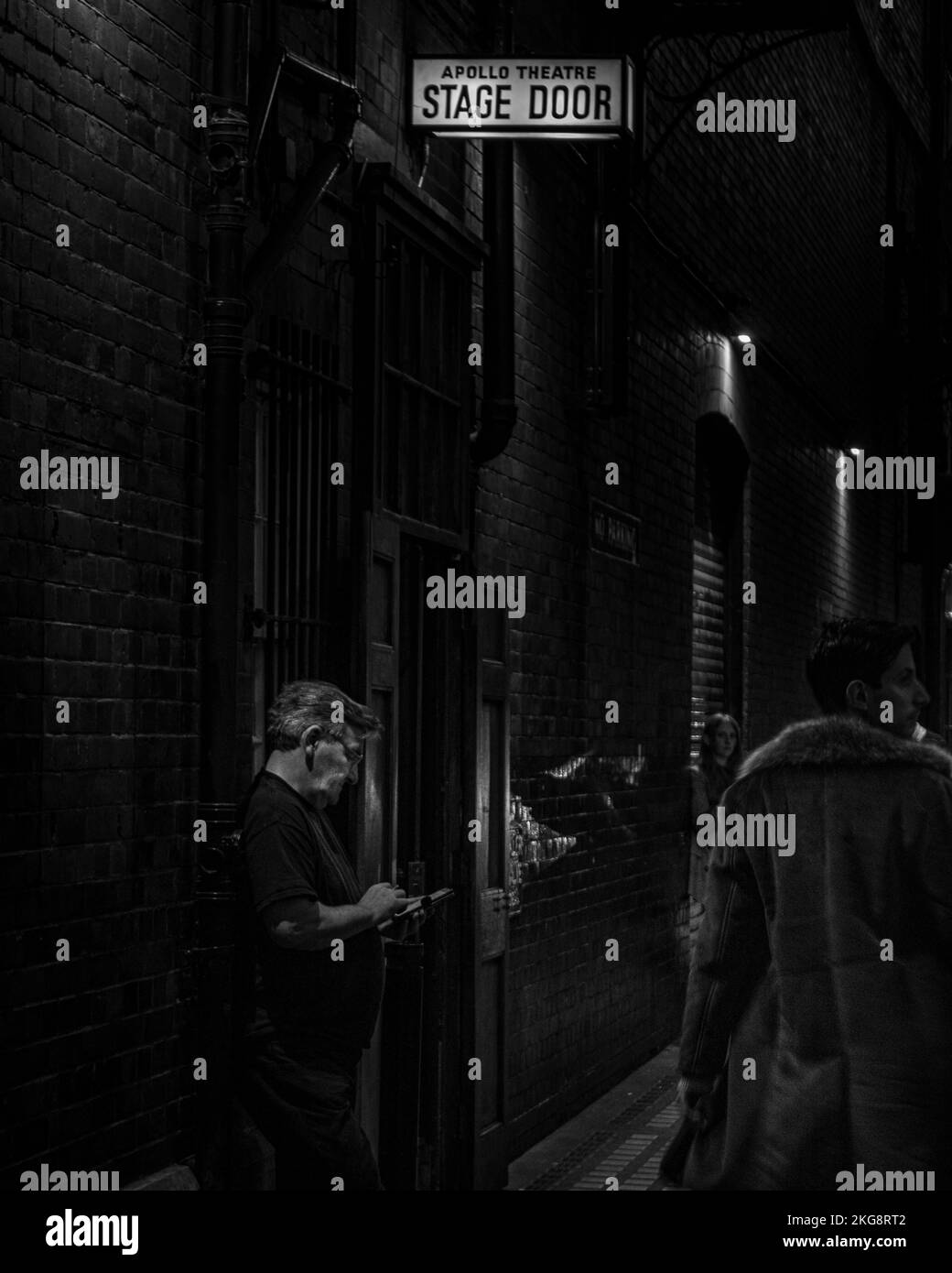 A black and white shot of a man smoking cigarette and scrolling on his phone underneath a stage door sign at the Apollo Theatre, London Stock Photo