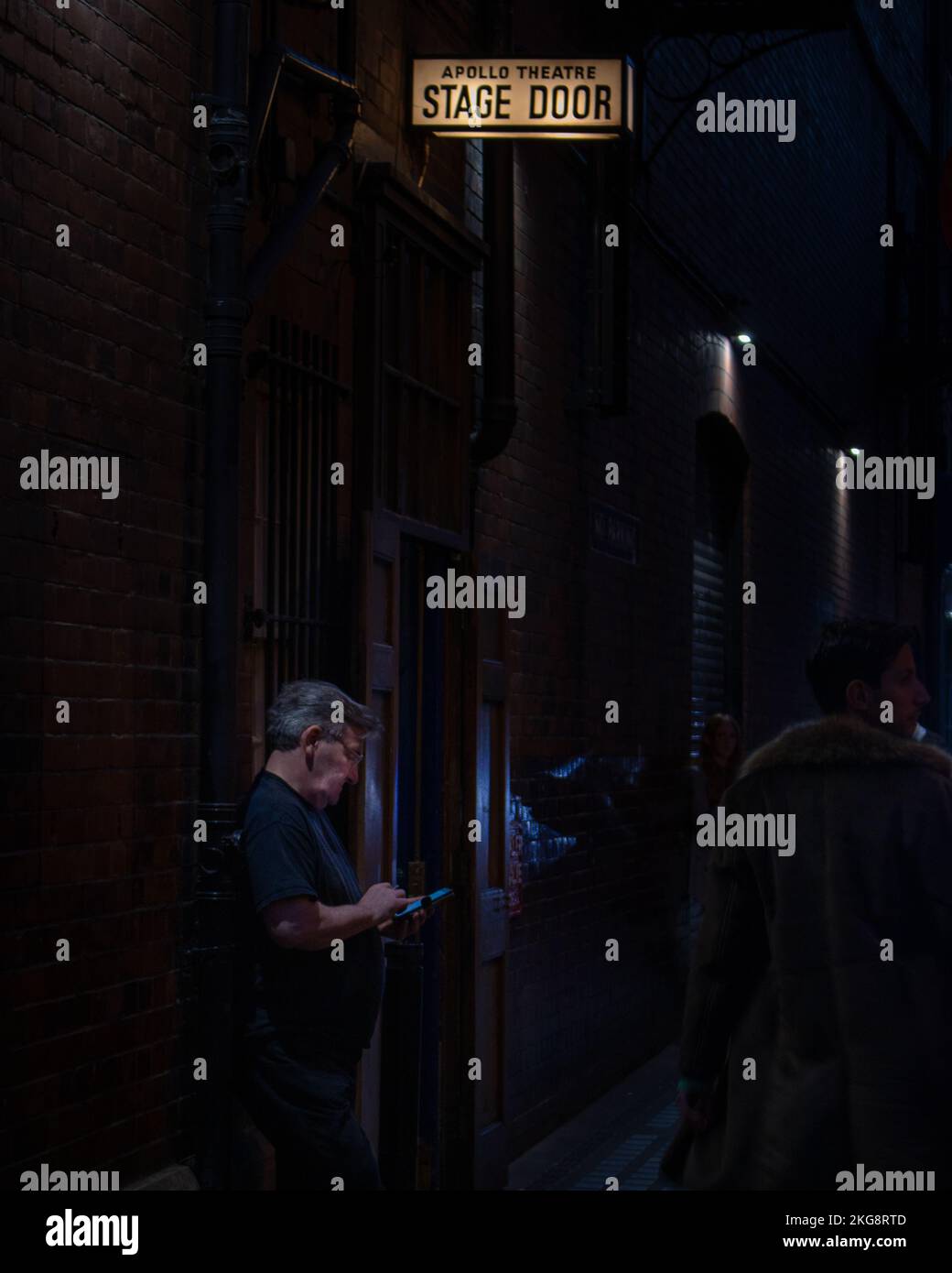 A low light photo of a man smoking a cigarette and checking his phone outside building underneath a stage door sign. Stock Photo