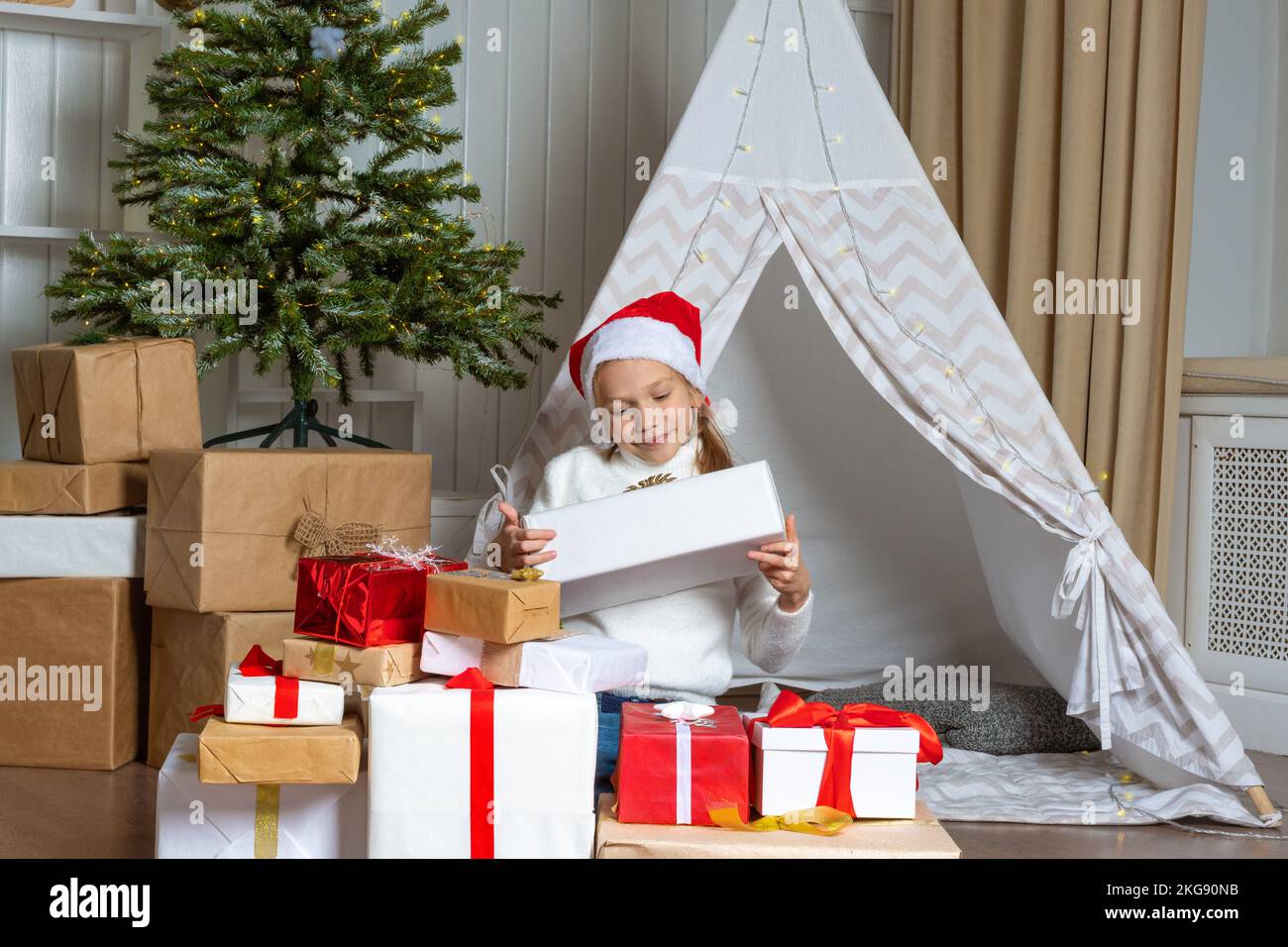 An emotional girl in a Santa hat is sitting on the floor next to a pile of her gifts in the nursery. The child is happy with gifts from Santa. The chi Stock Photo