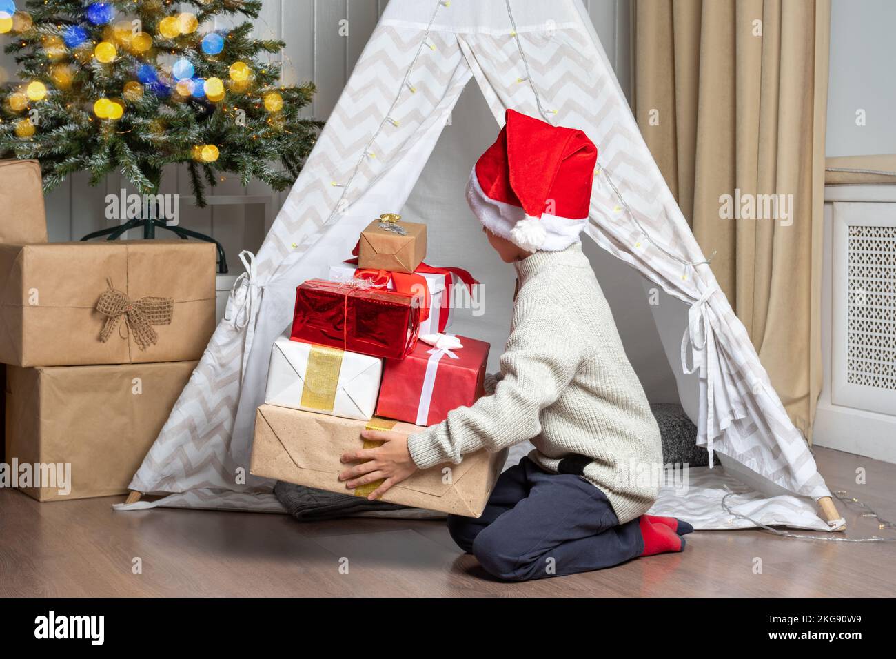 A pretty preschooler boy in a Santa hat hides his gifts in a children's tent wigwam in the nursery. The child wants to be alone with gifts. Kids Chris Stock Photo