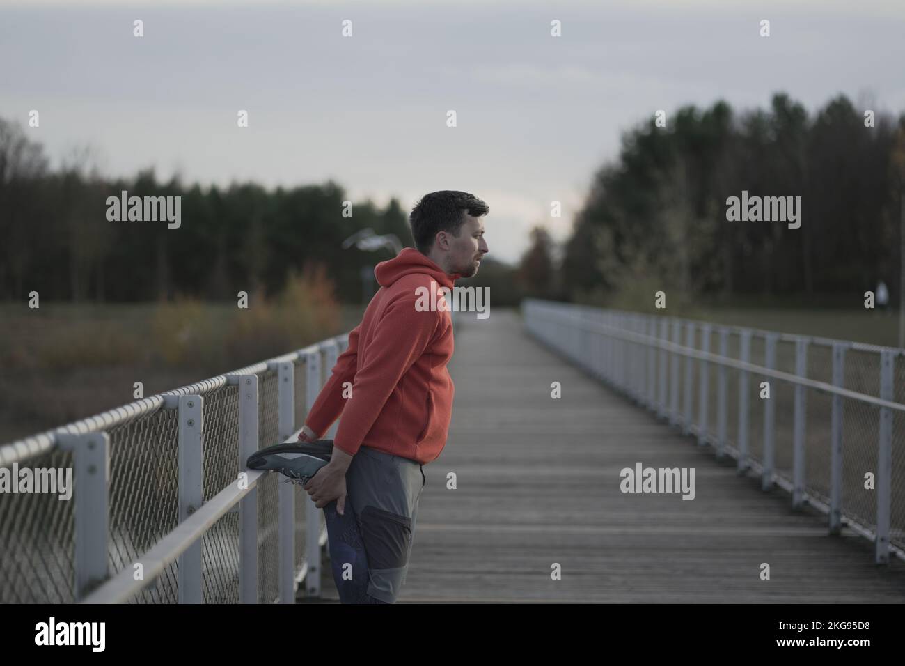 Man in orange and warm clothes warming up before jogging outdoors in cold autumn weather on wooden bridge. Runner stretching muscles leg and feet and Stock Photo