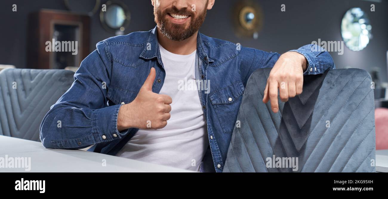 Unrecognyzable bearded man showing thumb up, while sitting at table in cafe. Crop view of smiling male in casual clothes gesturing like sign, while re Stock Photo