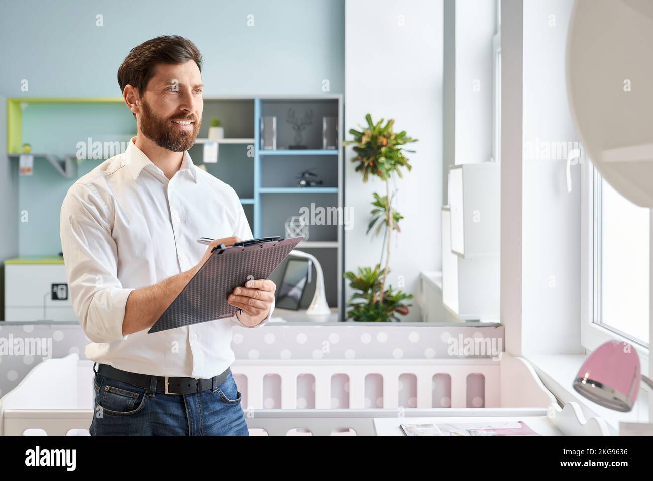 Successful sales manager standing with clip-on tablet in furniture store. Portrait of smiling retailer writing, compare prices, while looking at cupbo Stock Photo