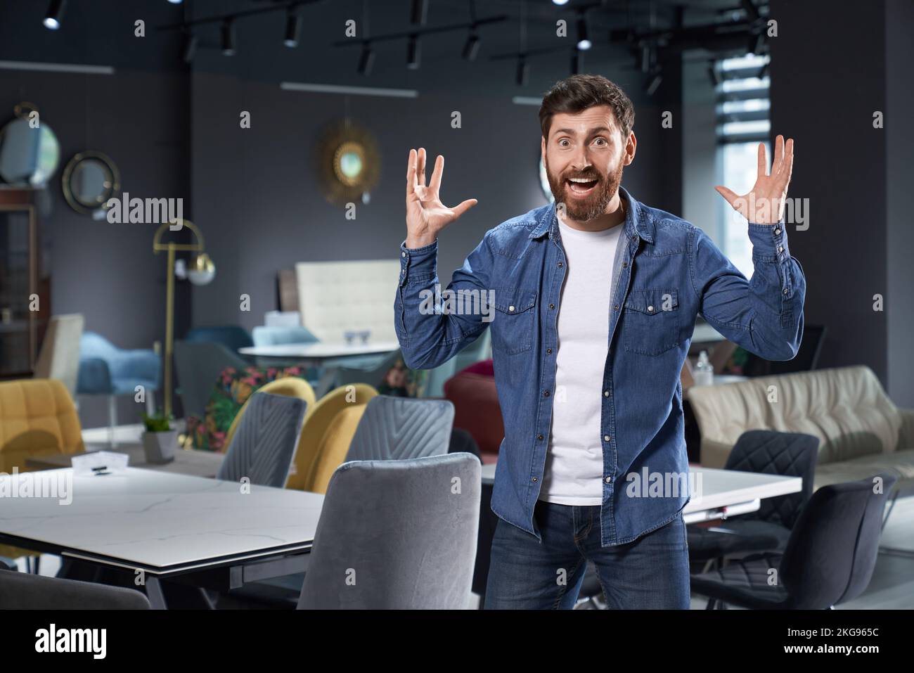Excited bearded customer raising hands from happiness, while standing in furniture store. Portrait of cheerful man in casual outfit feeling joy, while Stock Photo