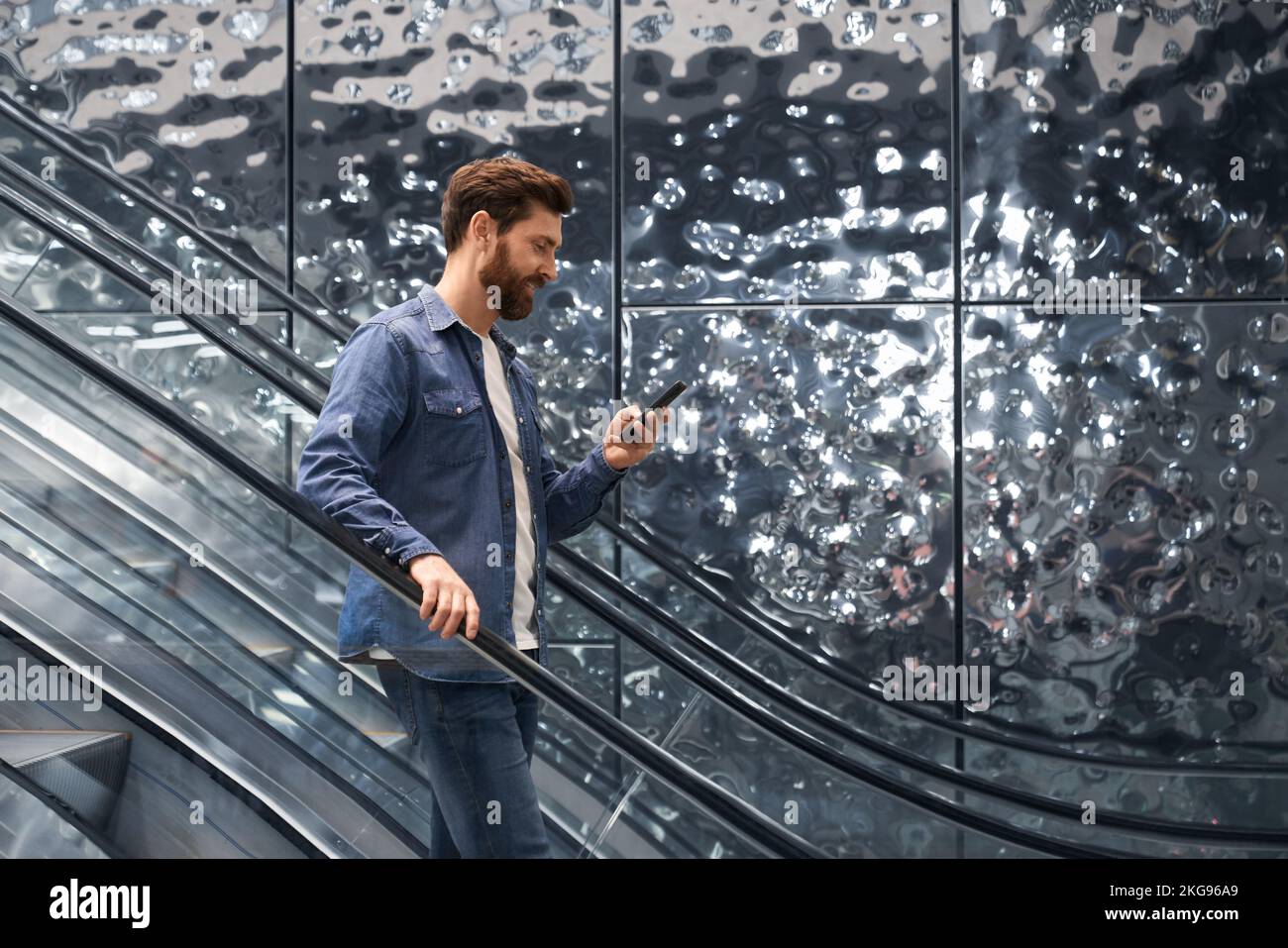 Smiling handsome man using smartphone, while going down on escalator of shopping mall. Side view of bearded male in denim jacket standing on moving st Stock Photo
