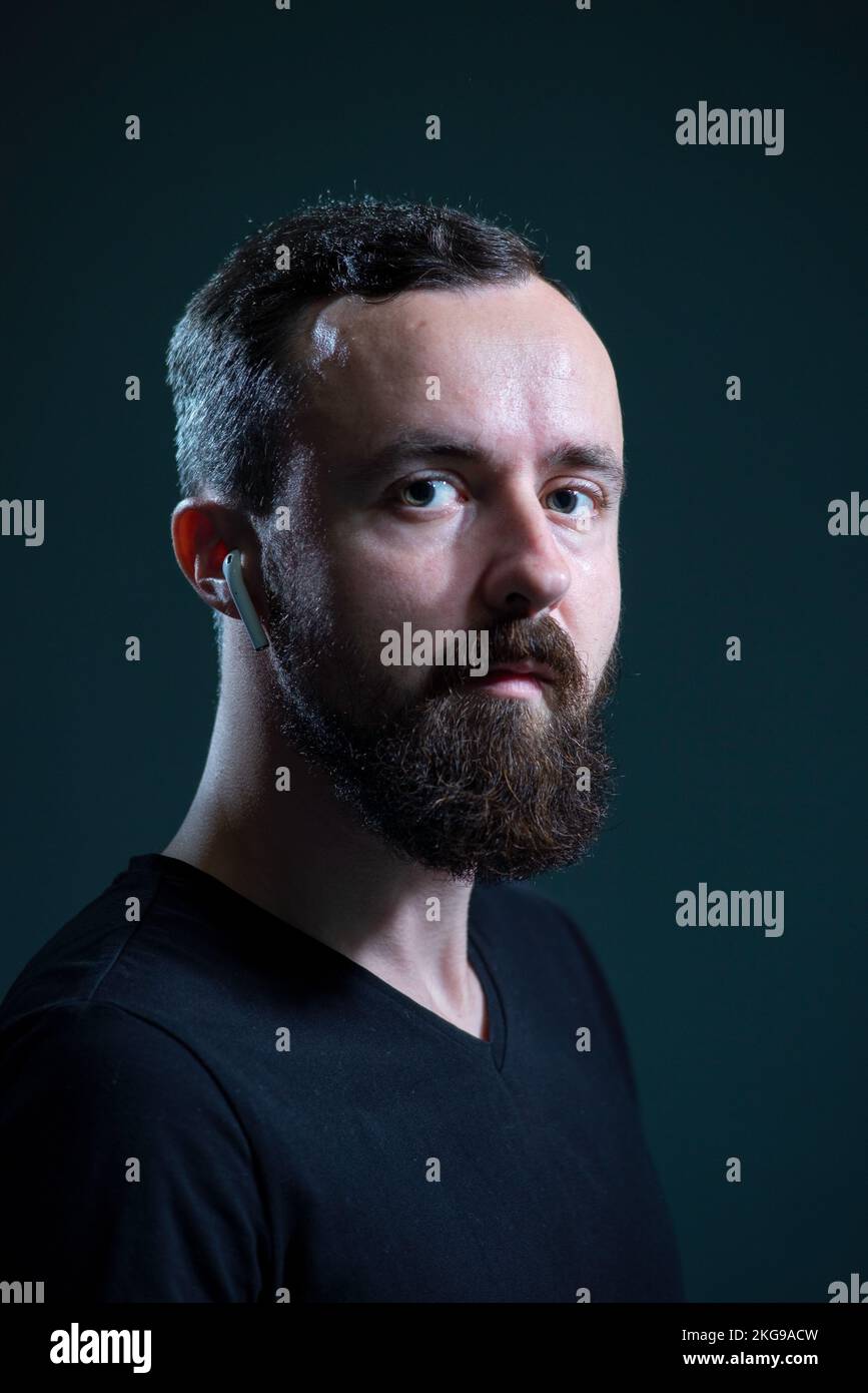 portrait of a bearded millennial guy in wireless headphones on a dark background. Stock Photo