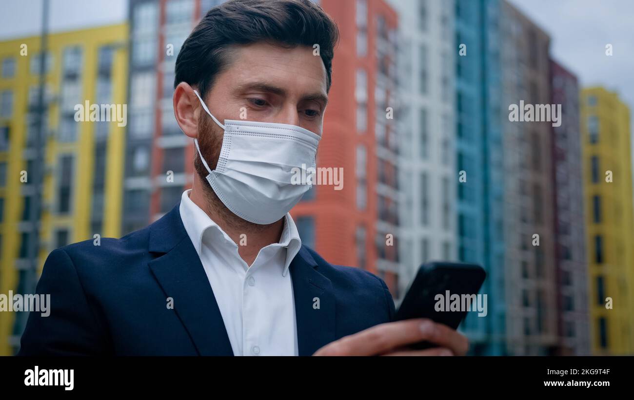 Male businessman in respiratory protection medical mask work on quarantine outdoor texting on smartphone at street professional chat with colleagues Stock Photo