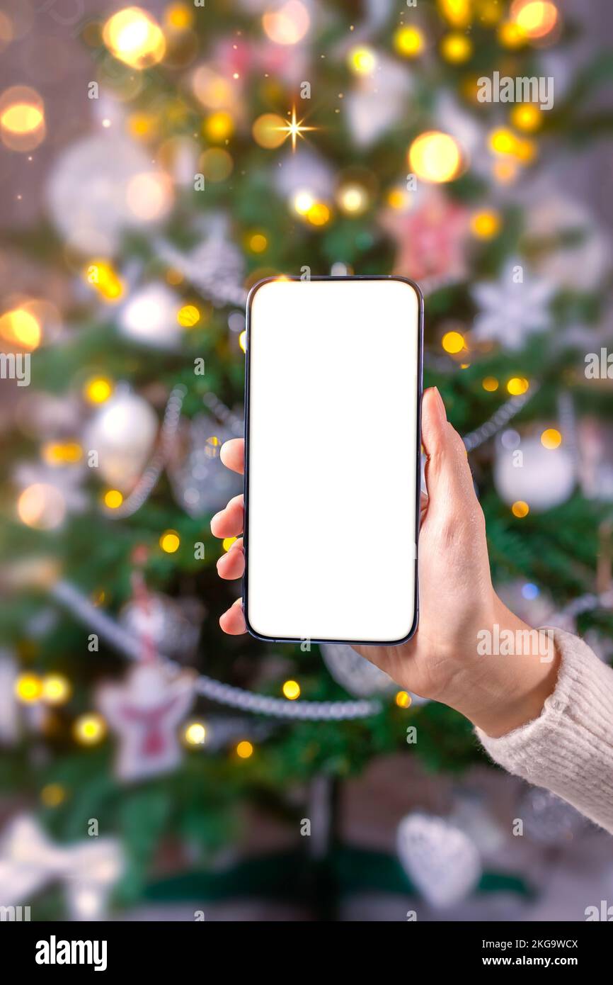 Close up smartphone with empty screen mockup on holiday background with Christmas tree, customer holding phone in hand, shopping online, purchasing gi Stock Photo