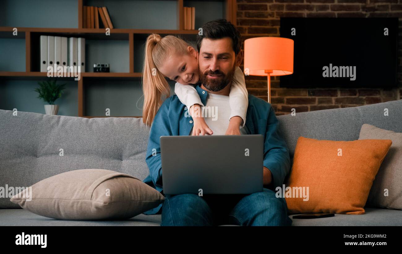 Cute active naughty girl noisy child plays with daddy hugs father distract parental attention disturbs parent from work on laptop at home single Stock Photo