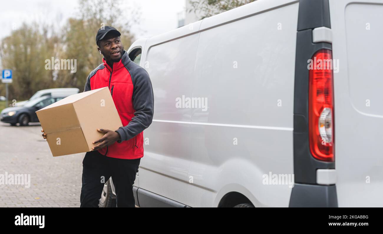 Strong focused Black man in vivid red-and-gray jacket holding one cardboard box in front of white delivery van. Package delivery concept. Blue-collar work concept. High quality photo Stock Photo