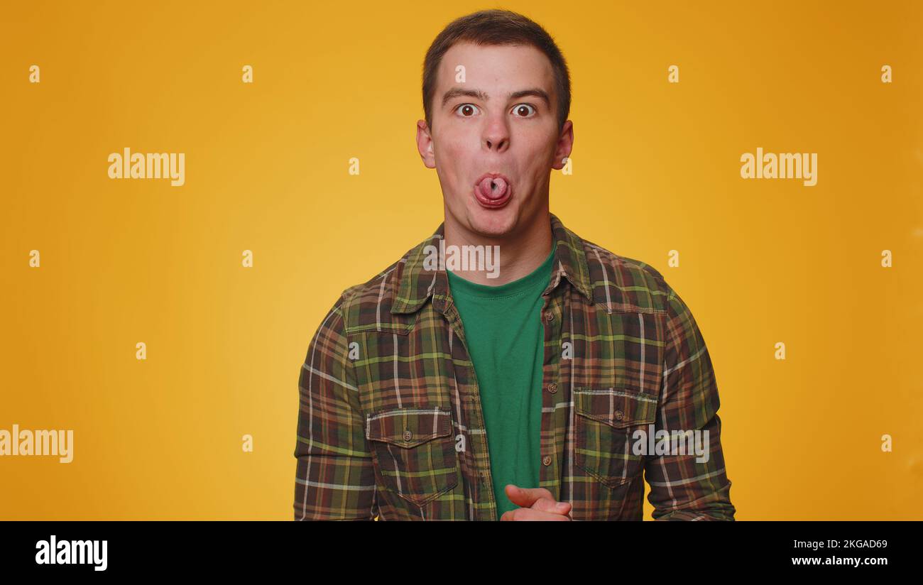 Cheerful funny man in casual green shirt showing tongue making faces at camera, fooling around, joking, aping with silly face, teasing. Young adult guy boy isolated alone on yellow studio background Stock Photo