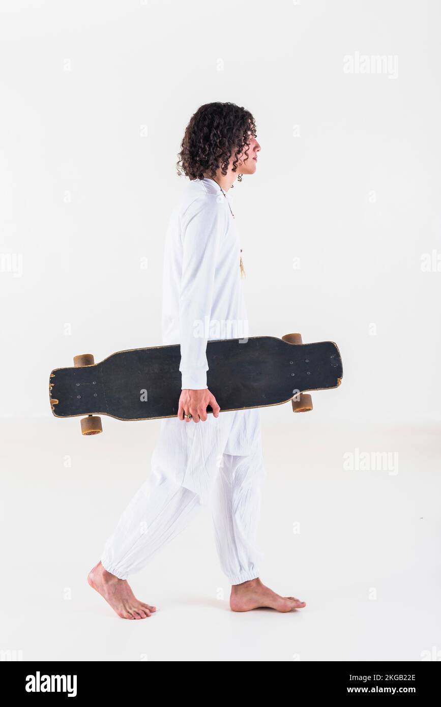 Side view of a young man with curly hair wearing white clothes and walking while holding a skate over white background Stock Photo