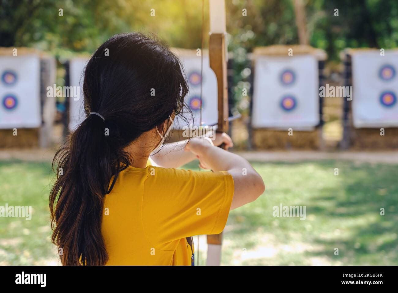 Back view of Asian girl wear eyeglasses with face mask aims archery bow and arrow to colorful