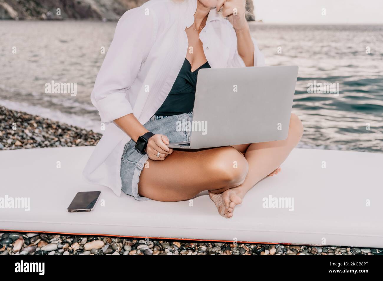 Digital nomad, Business woman working on laptop by the sea. Pretty lady typing on computer by the sea at sunset, makes a business transaction online Stock Photo