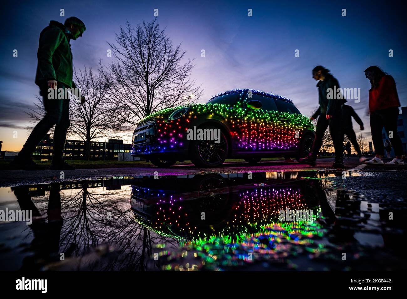People walk past an illuminated Mini car at the Mini Plant in Oxford. The driver of a Mini decorated in 3,000 twinkling lights hopes to 'bring little moments of joy to people's lives' this Christmas and raise over £10,000 for charity. Nicholas Martin, 33, is celebrating his fifth year behind the wheel of his Festive Mini but has gone to new lengths to create an illuminative show with thousands of app-controlled, battery-powered lights to generate different light patterns, messages, and animations. Picture date: Thursday November 17, 2022. Stock Photo