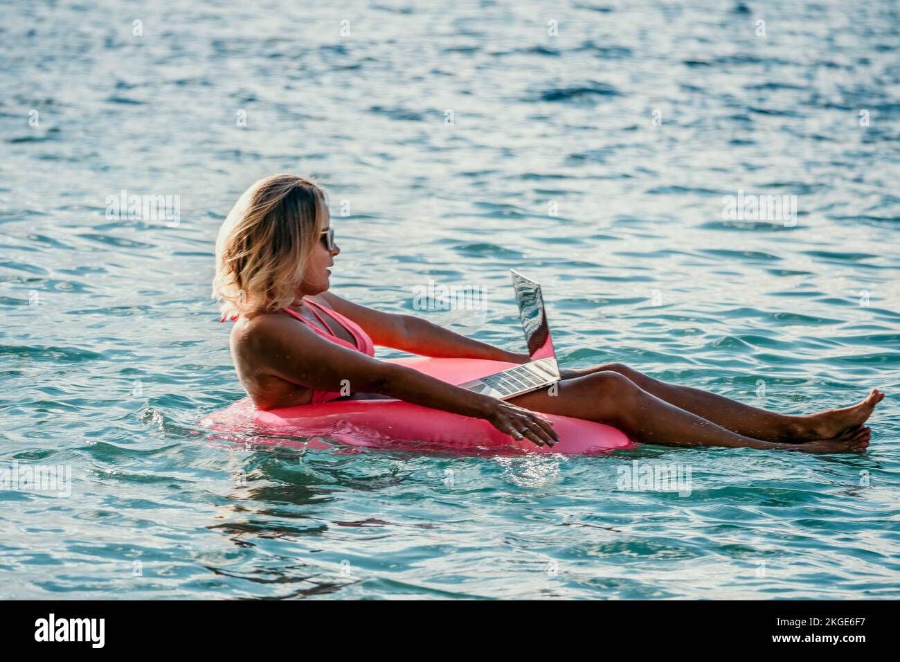 Woman works on laptop in sea. Freelancer, young blond woman in sunglases floating on an inflatable big pink donut with a laptop in the sea at sunset Stock Photo