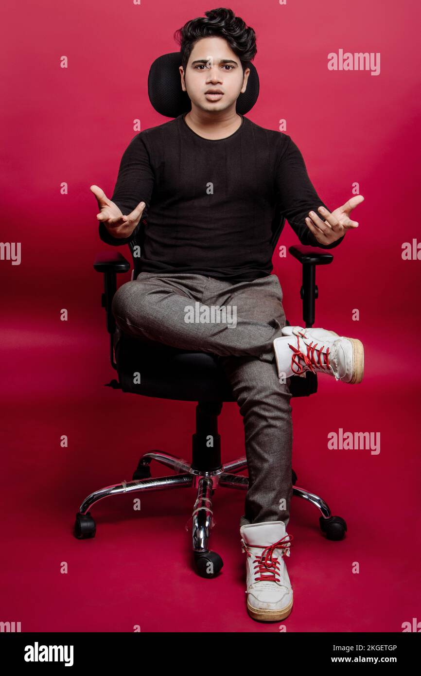 Frustrated young businessman manager sitting legs crossed on the chair looking at camera with hand gesture of questioning. Studio portrait over red Stock Photo