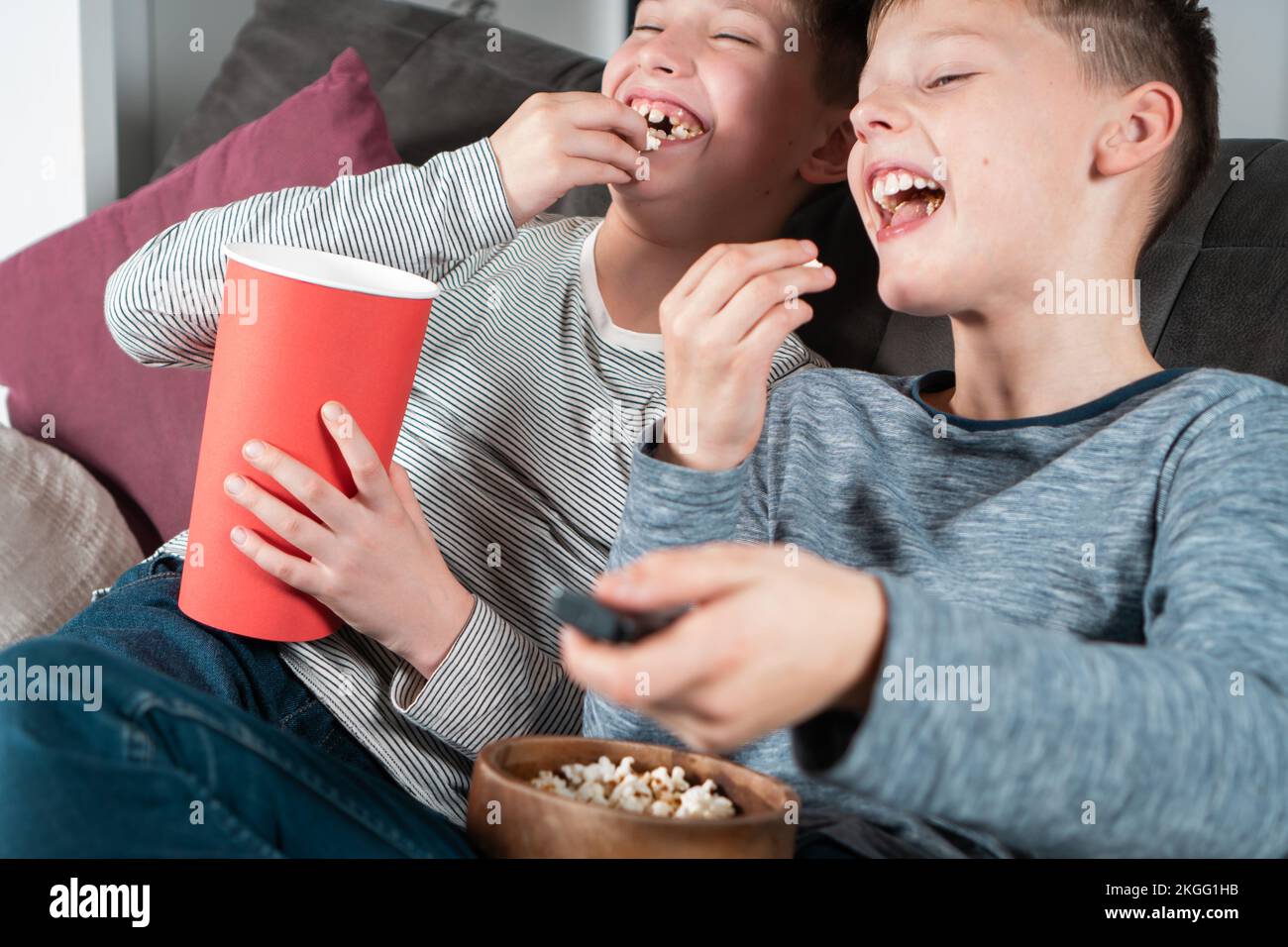 Portrait of two cheerful positive teenage boys sitting on sofa at home, watching funny movie comedy video cartoon on TV, eating popcorn from red Stock Photo