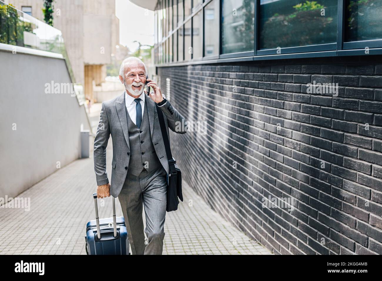Smiling senior businessman with suitcase talking on smart phone. Elderly professional wearing suit is carrying laptop bag. He is walking on footpath b Stock Photo