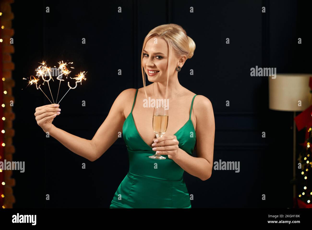 Front view of beautiful, blonde woman wearing green, seductive dress, smiling. Happy, young lady holding 2023 sparkles and glass of champagne. Concept Stock Photo