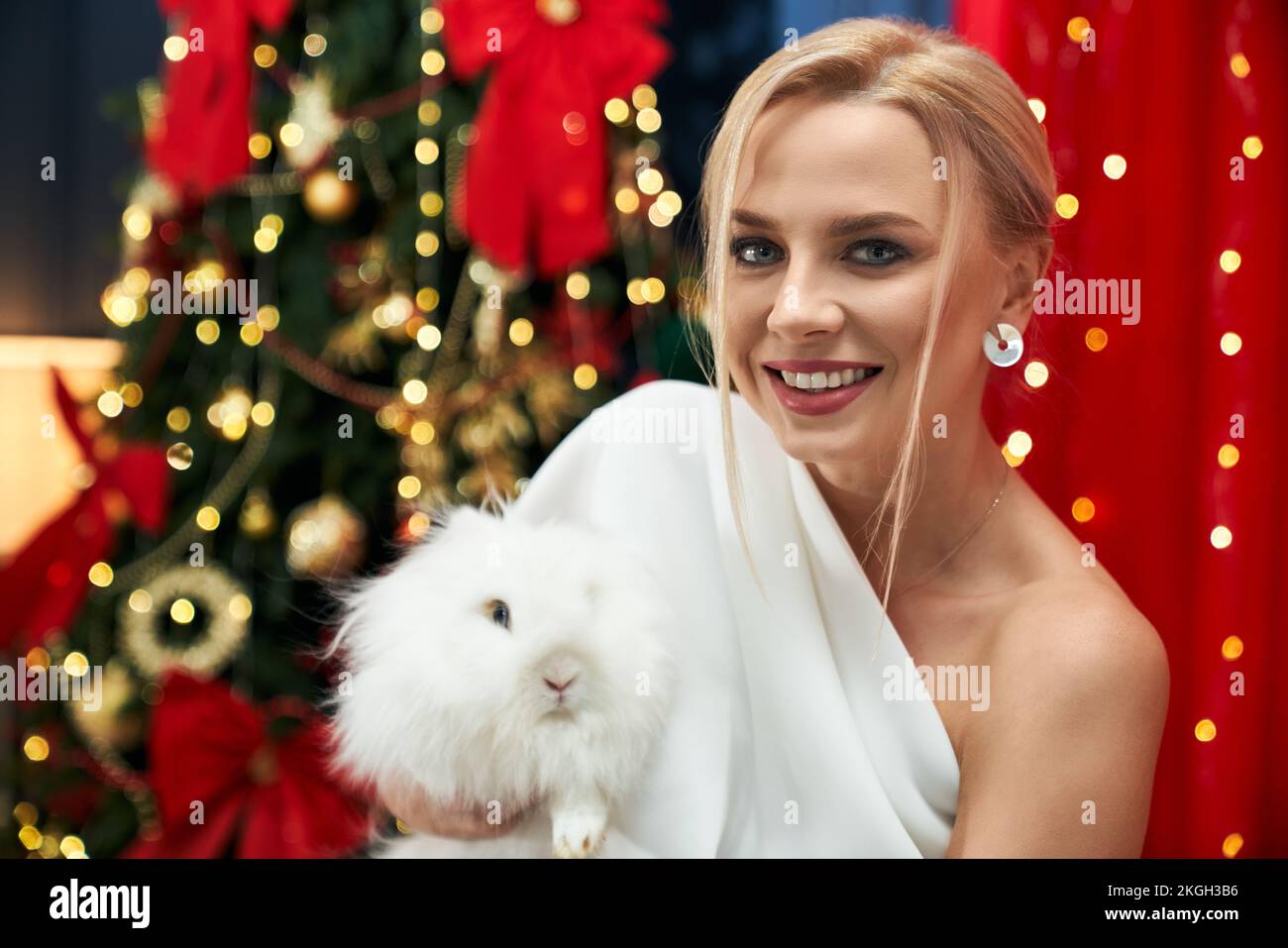 Front view of cheerful, happy lady holding white, furry rabbit. Beautiful, blonde woman wearing white dress, looking at camera, smiling. Concept of ho Stock Photo