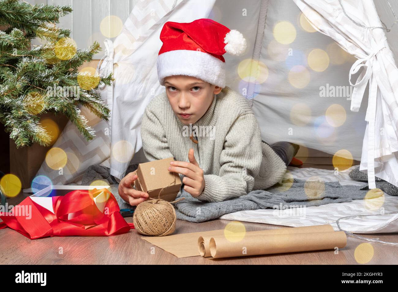 A child in a red Santa hat wraps Christmas gifts surprises in kraft paper with a rope, red ribbons. A boy lying in a children's tent packs gifts for t Stock Photo