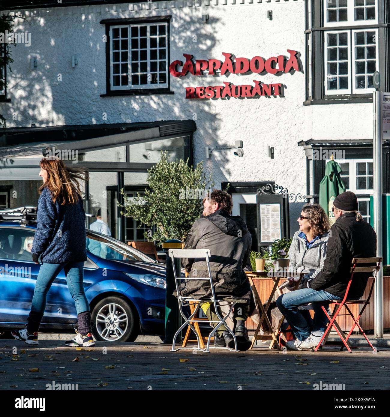 Epsom, Surrey, London UK, November 20 2022, Small Group Of People Sitting Outside Talking And Drinking Coffee With AWoman Walking Past Stock Photo