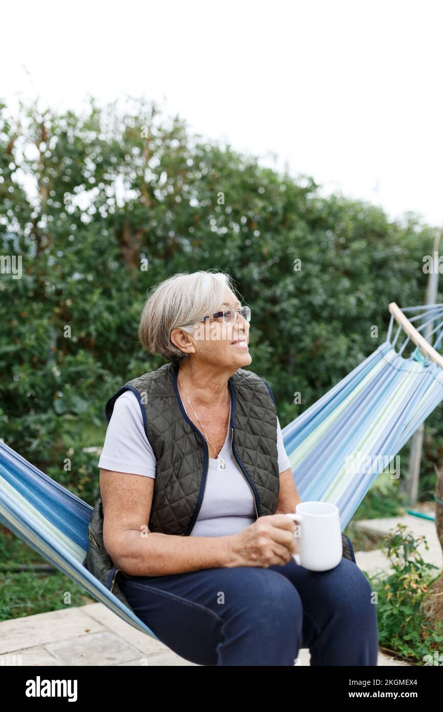 Beautiful senior blonde woman reading book and sitting in hammock in the garden Stock Photo